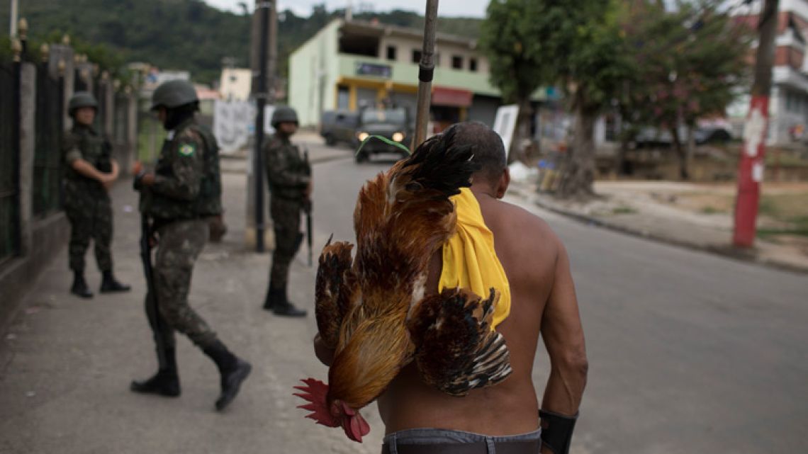 A man carries a rooster as walks on the sidewalk where soldiers stand guard in the Bateau Mouche slum of Rio de Janeiro, Brazil. The state's security ministry estimates that militias now control about a quarter of the sprawling state of Rio de Janeiro.In this May 19, 2018 photo, a man carries a rooster as walks on the sidewalk where soldiers stand guard in the Bateau Mouche slum of Rio de Janeiro, Brazil. The state's security ministry estimates that militias now control about a quarter of the sprawling state of Rio de Janeiro.