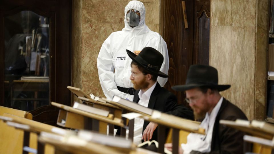 Un policía con protección a la pandemia espera que dos religiosos terminen de rezar en una sinagoga para detenerlos por violar las restricciones al coronavirus.