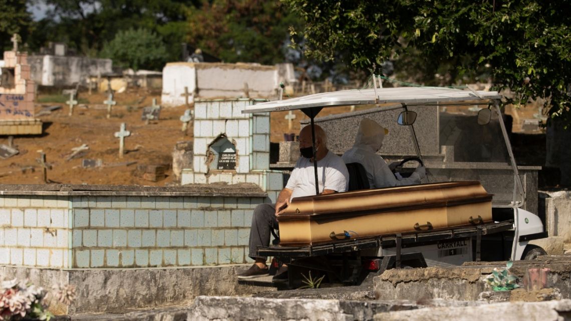 A man wearing a mask sits next the coffin of his mother as he's transported by a cemetery worker in a full protection suit to her burial site at the Nossa Senhora das Gracas cemetery in Duque de Caxias, Rio de Janeiro, Brazil, Monday, April 27, 2020. 