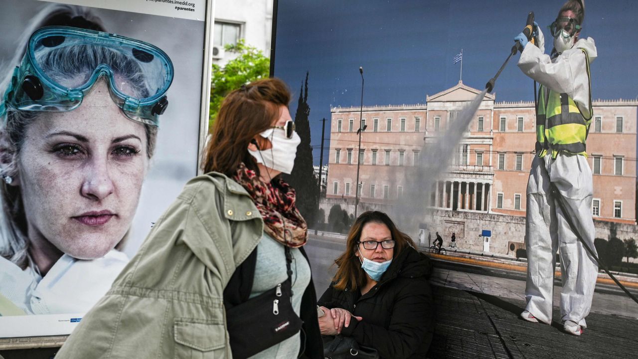 Una mujer (R) con una máscara protectora se sienta frente a un póster que muestra a una trabajadora limpiando el pavimento frente al Parlamento griego, en una parada de autobús en Atenas el 4 de mayo de 2020, el primer día de la relajación. restricciones de un bloqueo nacional contra la propagación del nuevo coronavirus, Covid-19. (Foto por ARIS MESSINIS / AFP) | Foto:AFP