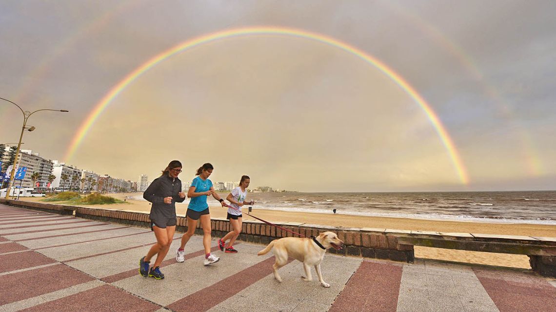 Women jog with a dog along Pocitos' Rambla as a double rainbow appears during sunset over Montevideo.