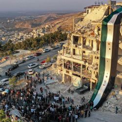 Esta fotografía muestra una vista aérea de sirios reunidos para una manifestación junto a las ruinas de un edificio que fue alcanzado en un bombardeo previo y adornado con una bandera gigante de la oposición siria, en la ciudad de Ariha en la rebelión. celebrada en el noroeste de la provincia de Idlib. | Foto:Omar HAJ KADOUR / AFP