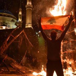 Un hombre libanés iza una bandera nacional mientras el 'puño de la Revolución', símbolo del levantamiento libanés de octubre de 2019, arde después de ser incendiado durante los enfrentamientos entre manifestantes antigubernamentales y partidarios del ex primer ministro Saad Hariri, en la plaza de los Mártires, en la capital, Beirut. | Foto:AFP