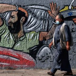 Un hombre palestino que lleva una máscara protectora pasa junto a un mural en el campo de refugiados de Nusseirat, en el centro de la Franja de Gaza, y expresa su apoyo al detenido administrativo bajo custodia israelí Maher al-Akhras, cuya salud se ha deteriorado mientras está en huelga de hambre durante casi 85 días. | Foto:Mohammed Abed / AFP