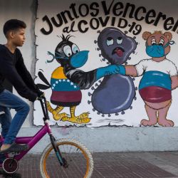 Un niño pasa en bicicleta junto a un mural que representa a un ave del grupo venezolano (icterus icterus) con la bandera de Venezuela y un oso con la bandera de Rusia, con máscaras y guantes, asfixiando una célula del virus COVID-19 y leyendo  | Foto:Cristian Hernandez / AFP