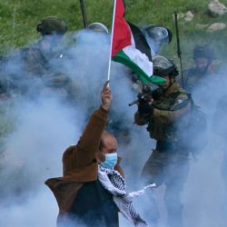 Un manifestante palestino iza una bandera nacional mientras un miembro de las fuerzas israelíes apunta su arma, en medio del humo de los gases lacrimógenos durante una manifestación contra el establecimiento de puestos de avanzada israelíes en sus tierras, en Beit Dajan, al este de Nablus en la Cisjordania ocupada. | Foto:Jaafar Ashtiyeh / AFP