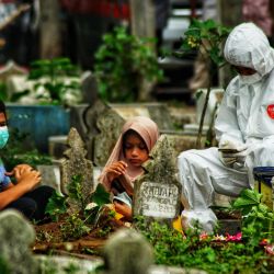 Un miembro de la familia con traje de equipo de protección personal (PPE) junto con dos niños rezan junto a la tumba de una víctima del coronavirus Covid-19 que acaba de ser enterrada, mientras los miembros de la familia que desafían los protocolos de salud se reúnen cerca en un cementerio en Lhokseumawe, Aceh. | Foto:Azwar Ipank / AFP