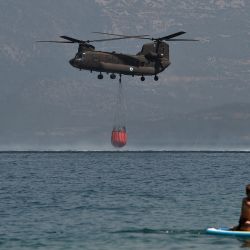 Un helicóptero Ch-47D Chinook es observado por un practicante de paddleboard mientras se llena de agua durante la lucha contra el fuego cerca de la playa de Lambiri en Patras. | Foto:Louisa Gouliamaki / AFP