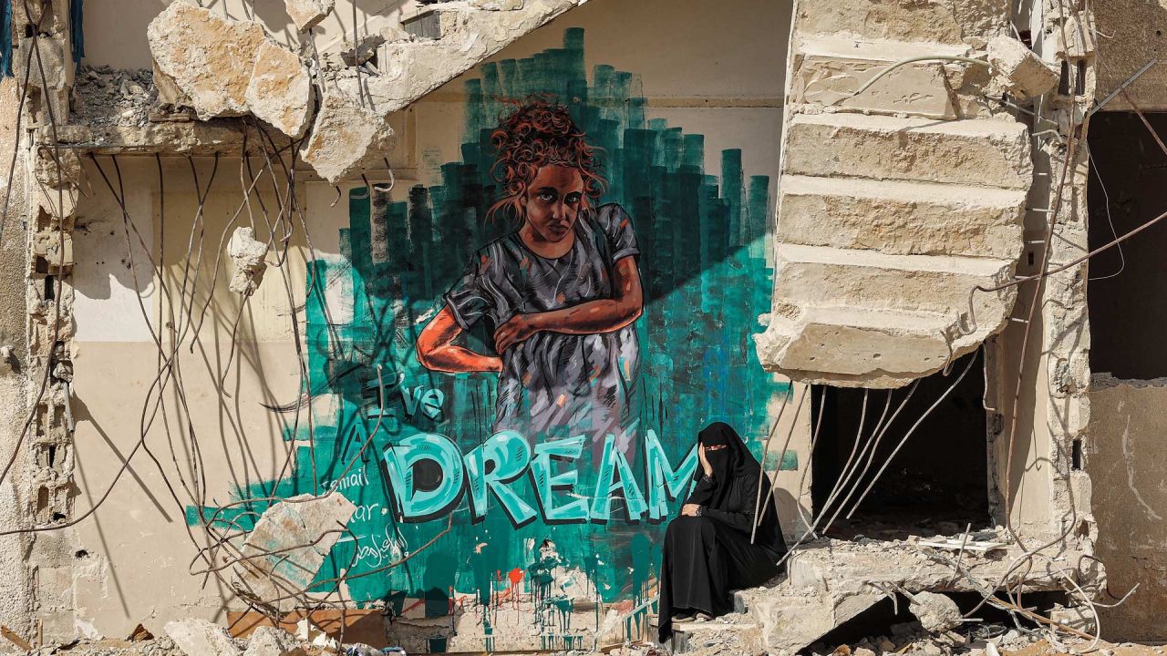 Una mujer se sienta junto a los escalones rotos de un edificio parcialmente derrumbado y destruido por los bombardeos durante el conflicto de mayo de 2021 entre Hamás e Israel, junto a un mural de grafitis que representa a una niña con una leyenda en inglés debajo que dice  | Foto:Mohammed Abed / AFP