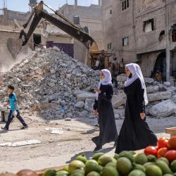 Unas mujeres pasan por delante de un puesto de verduras, mientras unos trabajadores limpian los escombros de un edificio destruido por los bombardeos israelíes el pasado mayo, en Rafah, en el sur de la Franja de Gaza. | Foto:Said Khatib / AFP