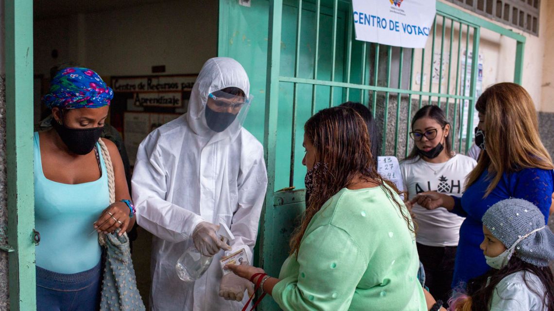 An electoral staffer wearing a protective biosecurity suit sprays disinfectant to a voter hands at the entrance of a polling station during the regional and municipal elections in Caracas, on November 21, 2021. 