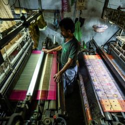 En esta fotografía un trabajador utiliza un telar mecánico para tejer un sari de seda Banarasi en un taller de Benarés, India. - Los tejedores manuales de Varanasi han cultivado una reputación de excelencia a lo largo de los siglos, especializándose en patrones intrincados, diseños florales y brocados dorados. | Foto:MONEY SHARMA / AFP