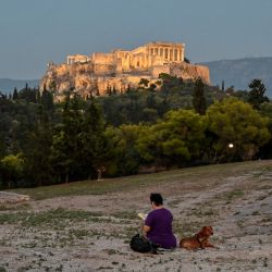 Una mujer lee un libro junto a su perro en la colina de Pnyx en Atenas, frente a la Acrópolis illumanada. | Foto:LOUISA GOULIAMAKI / AFP