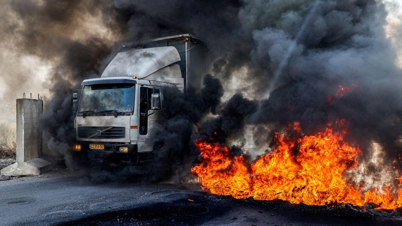 Un camión que se dirige a Nablus pasa por delante de unos neumáticos en llamas en el puesto de control de Hawara, al sur de la ciudad palestina en la Cisjordania ocupada, que fueron incendiados por manifestantes palestinos que exigían la reapertura de las carreteras en torno a la ciudad cerrada de Nablus, que ha estado bajo bloqueo militar desde el 11 de octubre después de que hombres armados palestinos mataran a un soldado israelí cerca del asentamiento de Shavei Shomron. | Foto:JAAFAR ASHTIYEH / AFP