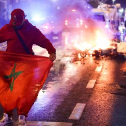 Un aficionado sostiene una bandera marroquí mientras unos patinetes eléctricos arden por detrás en la banda de la retransmisión en directo del partido de fútbol del Grupo F de la Copa del Mundo de Qatar 2022 entre Bélgica y Marruecos, en Bruselas. | Foto:KENZO TRIBOUILLARD / AFP