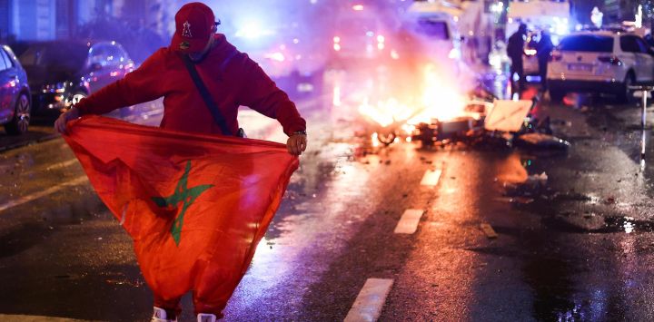 Un aficionado sostiene una bandera marroquí mientras unos patinetes eléctricos arden por detrás en la banda de la retransmisión en directo del partido de fútbol del Grupo F de la Copa del Mundo de Qatar 2022 entre Bélgica y Marruecos, en Bruselas.