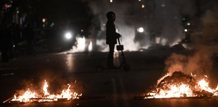 Esta fotografía muestra a una mujer pasando junto a las barricadas en llamas colocadas por miembros de la comunidad romaní en Grecia frente al hospital donde un adolescente romaní recibe tratamiento tras ser tiroteado por la policía, en Salónica.
