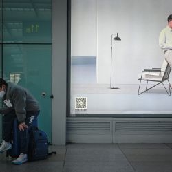 Un hombre sentado en su equipaje utiliza su teléfono móvil delante de un anuncio en la estación de tren sur de Pekín, en Pekín, China. | Foto:Wang Zhao / AFP