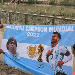 Un hombre y una mujer despliegan una bandera de Argentina que representa a los delanteros Diego Maradona y Lionel Messi con el texto en español "Argentina campeón mundial 2022" mientras peregrinos cristianos se adentran en el río Jordán en el sitio bautismal de Qasr al-Yahud. Foto de HAZEM BADER / AFP | Foto:AFP