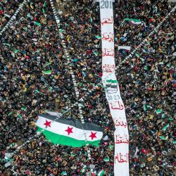 Esta vista aérea muestra a personas portando una bandera gigante de la oposición siria antes de ser izada en un mástil, durante una manifestación para conmemorar el 12º aniversario del inicio del levantamiento contra el presidente sirio Bashar al Assad y su gobierno, en la ciudad noroccidental de Idlib, controlada por los rebeldes. | Foto:OMAR HAJ KADOUR / AFP