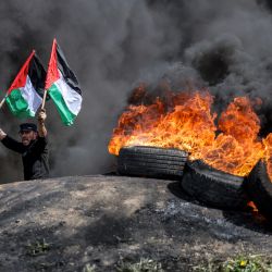Un hombre sostiene banderas palestinas mientras permanece junto a los humos de neumáticos en llamas durante una manifestación en la frontera con Israel, al este de la ciudad de Gaza. | Foto:MAHMUD HAMS / AFP