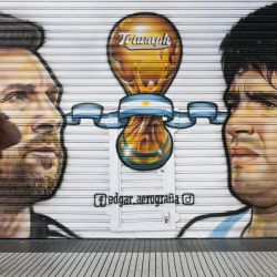 Una mujer pasa por delante de una tienda cerrada donde están pintadas las imágenes de los capitanes de la selección argentina de fútbol, Lionel Messi (campeón del mundo en Qatar 2022) y el fallecido astro del fútbol Diego Maradona (campeón del mundo en México 1986), en Buenos Aires. | Foto:JUAN MABROMATA / AFP