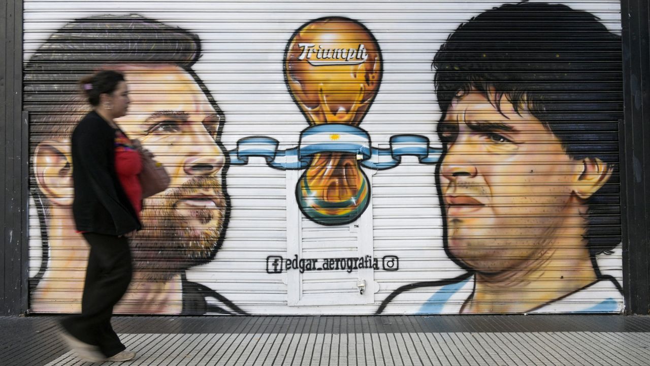 Una mujer pasa por delante de una tienda cerrada donde están pintadas las imágenes de los capitanes de la selección argentina de fútbol, Lionel Messi (campeón del mundo en Qatar 2022) y el fallecido astro del fútbol Diego Maradona (campeón del mundo en México 1986), en Buenos Aires. | Foto:JUAN MABROMATA / AFP