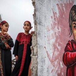 Niños palestinos posan junto a un mural durante una exposición de arte mixto titulada "La ocupación mata a la infancia" que tiene lugar entre los escombros de las casas destruidas por los ataques aéreos israelíes durante el conflicto de mayo de 2023 en Deir al-Balah, en el centro de la Franja de Gaza. | Foto:MOHAMMED ABED / AFP