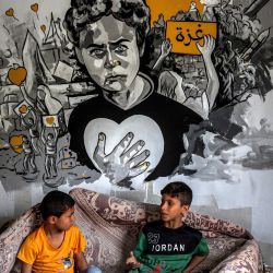 Niños palestinos sentados bajo un mural durante una exposición de arte mixto titulada "La ocupación mata a la infancia" que tiene lugar entre los escombros de las casas destruidas por los ataques aéreos israelíes durante el conflicto de mayo de 2023 en Deir al-Balah, en el centro de la Franja de Gaza. | Foto:MOHAMMED ABED / AFP