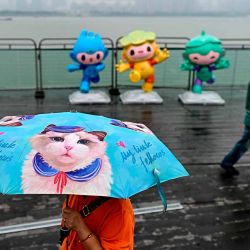 Una mujer que lleva un paraguas pasa junto a las mascotas de los Juegos Asiáticos, Chenchen. Foto de MANAN VATSYAYANA / AFP  | Foto:AFP