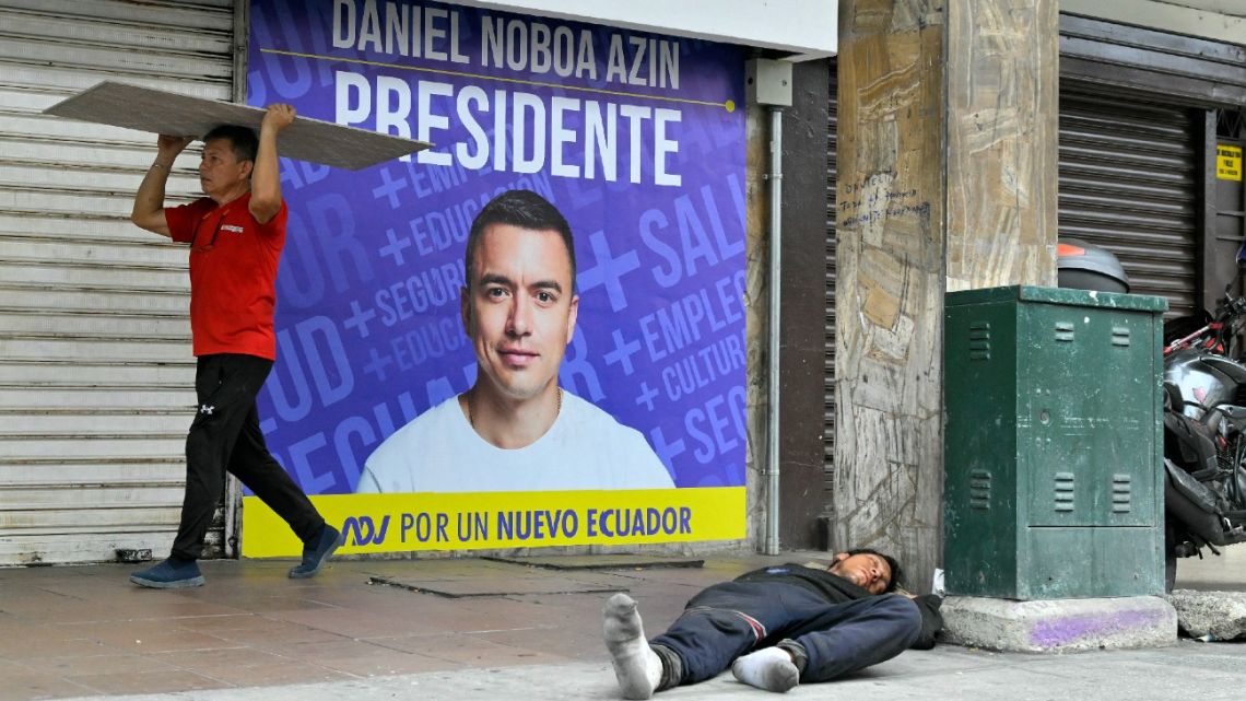 A man sleeps in the street while another person walks in front of advertisement for Ecuador's presidential candidate of the National Democratic Action Party, Daniel Noboa, in Guayaquil, Ecuador, on October 13, 2023. 