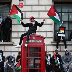 Un manifestante ondea banderas palestinas sentado en una cabina telefónica roja en Whitehall durante la "Marcha por Palestina" en Londres, para pedir un alto el fuego en el conflicto entre Israel y Hamás. | Foto:HENRY NICHOLLS / AFP