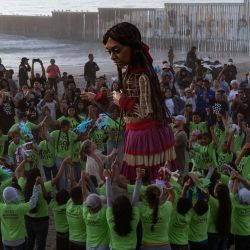 Vista aérea de la pequeña Amal, una marioneta de 12 pies de altura que representa a un niño refugiado sirio de diez años, durante un baile con niños migrantes cerca del muro fronterizo entre Estados Unidos y México en Playas de Tijuana, estado de Baja California. La pequeña Amal visita Tijuana en el marco de un viaje por México para crear conciencia sobre la difícil situación de los refugiados y migrantes en todo el mundo. | Foto:Guillermo Arias/AFP