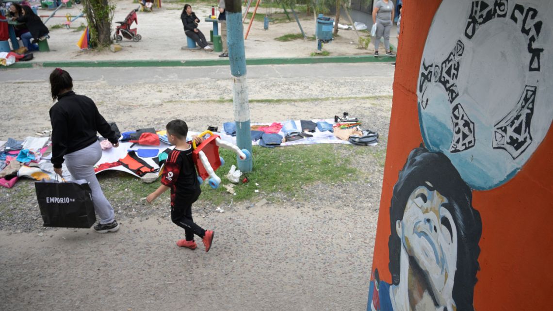 People pass by used clothes for barter or sale on a sidewalk in Villa Fiorito, Lomas de Zamora on November 13, 2023 where a pedestrian bridge is decorated with portraits of late football star Diego Maradona. 
