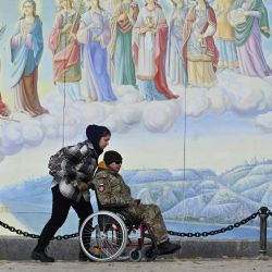 Una niña lleva una silla de ruedas con un militar discapacitado frente a los frescos de la Catedral de las Cúpulas Doradas de San Miguel en Kiev, en medio de la invasión rusa de Ucrania. | Foto:SERGEI SUPINSKY / AFP