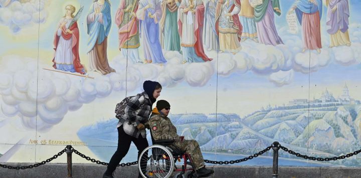 Una niña lleva una silla de ruedas con un militar discapacitado frente a los frescos de la Catedral de las Cúpulas Doradas de San Miguel en Kiev, en medio de la invasión rusa de Ucrania.