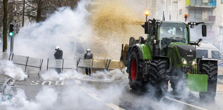 Un agricultor arroja heno de su tractor a los agentes de la policía antidisturbios belgas mientras el gas se eleva de los botes de gas lacrimógeno durante una protesta convocada por las organizaciones de agricultores "Federation Unie de Groupements d'Eleveurs et d'Agriculteurs" (FUGEA), Boerenforum y MAP, en respuesta al Consejo Europeo de Agricultura, en Bruselas.
