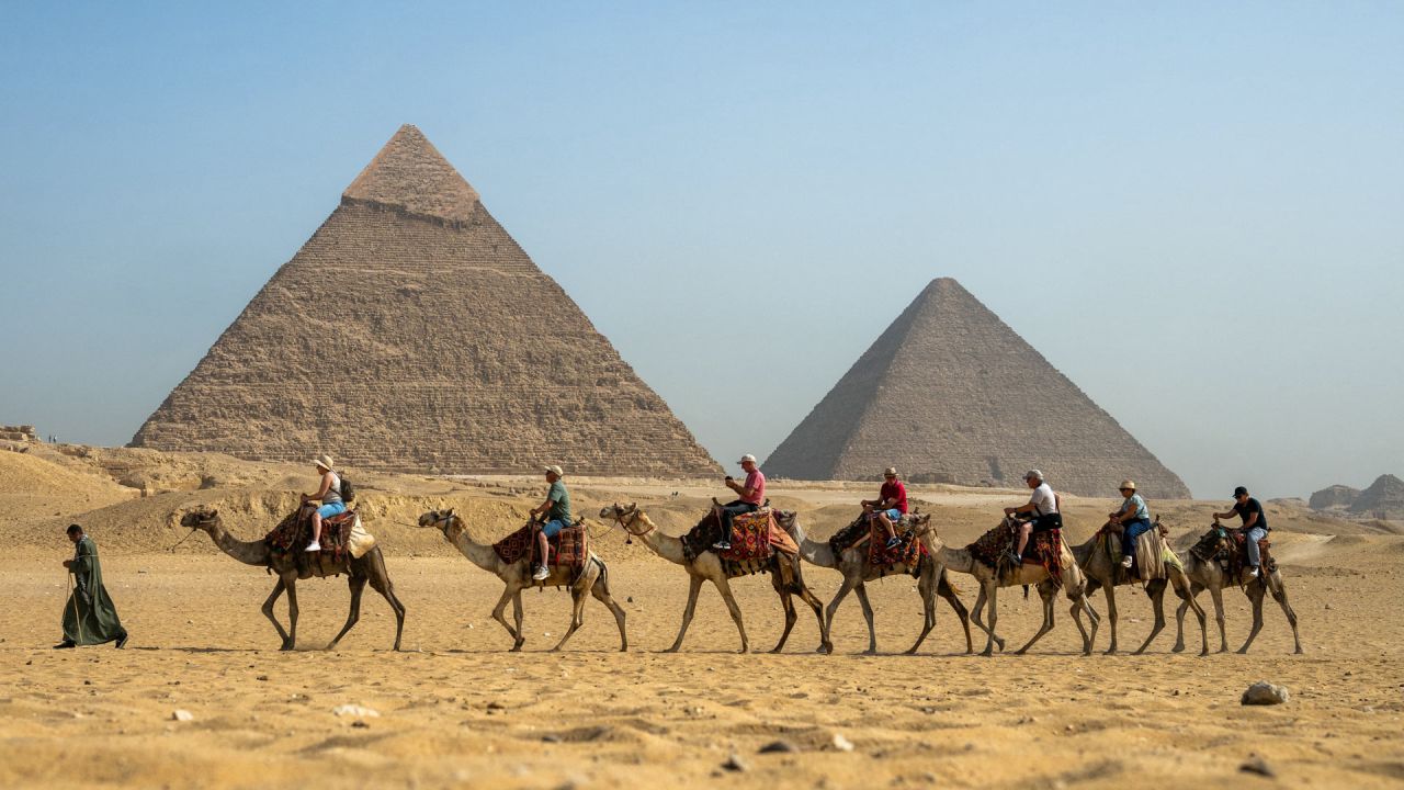 Un guía tira de un camello mientras los turistas pasan en fila pasando por la Gran Pirámide de Keops y la Pirámide de Khafre en la Necrópolis de las Pirámides de Giza en las afueras de Giza. | Foto:Joya Samad / AFP