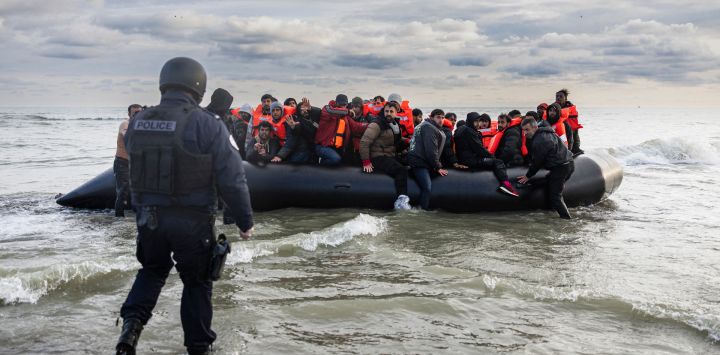 Los inmigrantes reaccionan mientras un oficial de policía francés está listo para perforar el barco del contrabandista con un cuchillo para evitar que los inmigrantes se embarquen en un intento de cruzar el Canal de la Mancha en la playa de Gravelines, cerca de Dunkerque, al norte de Francia.