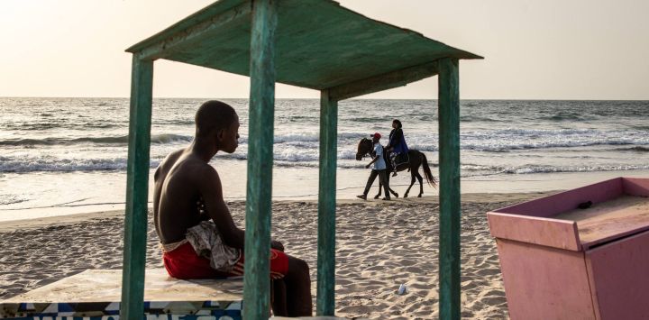 Un joven mira a una mujer dando un paseo a caballo por una playa en Banjul, Gambia.