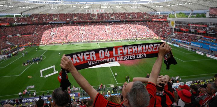 Un fan del Bayer Leverkusen muestra una bufanda con la inscripción 'Bayer 04 Leverkusen - Campeones alemanes 2024' en el estadio BayArena en Leverkusen, Alemania occidental, mientras los jugadores y aficionados del Bayer Leverkusen celebran después del partido de fútbol de la primera división alemana de la Bundesliga. entre Bayer 04 Leverkusen y FC Augsburgo. El Bayer Leverkusen se ha convertido en el primer equipo en la historia de la Bundesliga en pasar una temporada invicto después de una victoria en casa por 2-1 sobre el Augsburgo, ampliando su racha de temporada a 51 partidos sin derrotas.