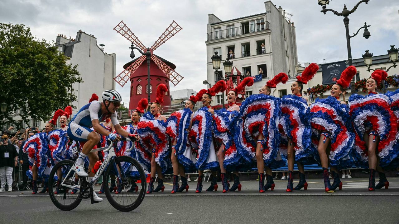 El ciclista francés Valentin Madouas pasa en bicicleta junto a bailarines del Moulin Rouge que realizan el cancán durante la carrera de ciclismo en ruta masculina durante los Juegos Olímpicos de París 2024 en París. | Foto:MAURO PIMENTEL / AFP