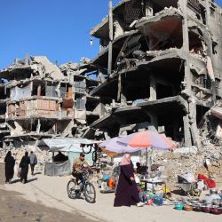 Un hombre pasa en bicicleta junto a vendedores que exhiben sus productos en el exterior de edificios gravemente dañados en un campamento que alberga a personas desplazadas por el conflicto en Jabalia, en el norte de la Franja de Gaza, en medio de la guerra en curso en el territorio palestino entre Israel y Hamás. | Foto:OMAR AL-QATTAA / AFP