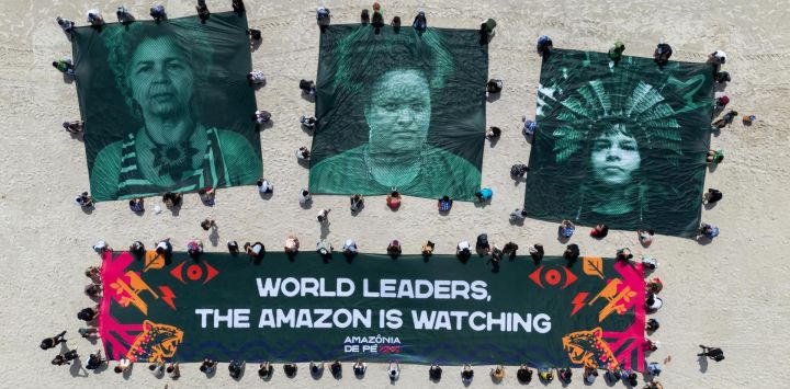 Activistas ambientales muestran una pancarta gigante que dice: "Líderes mundiales, la Amazonia está mirando" y fotos de los activistas ambientales brasileños Angela Mendes, Kelly Araujo y Txai Surui en la playa de Botafogo con el cerro Pan de Azúcar al fondo en Río de Janeiro, durante la Cumbre del G20.