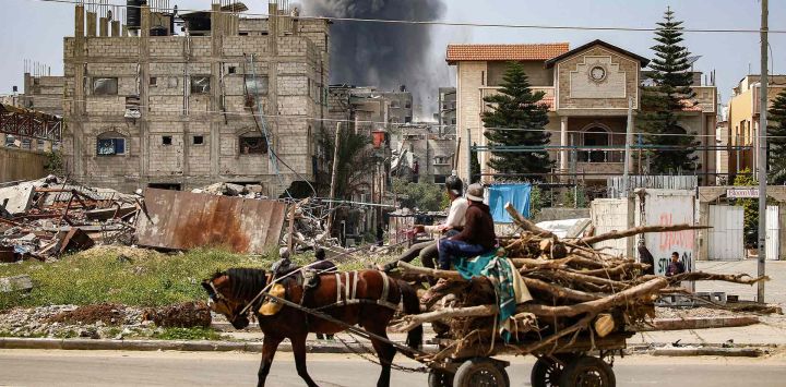 Hombres en una carreta tirada por caballos, observan cómo una columna de humo se eleva desde un edificio alcanzado por un bombardeo israelí. Foto de Eyad BABA / AFP
