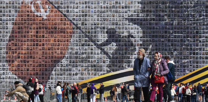 La gente pasa junto a una pancarta gigante de "Izando la Bandera sobre el Reichstag", que representa a un soldado del Ejército Rojo izando la Bandera Roja de la URSS en lo alto del edificio del Reichstag en Berlín. Esta pancarta está hecha con 4500 retratos de veteranos de la Segunda Guerra Mundial y lleva el mensaje "Nadie es olvidado, nada es olvidado".