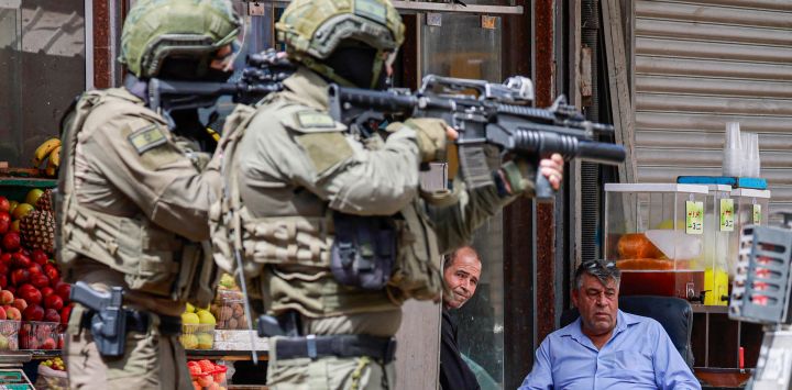 Soldados del ejército israelí apuntan con sus rifles mientras palestinos sentados frente a una tienda observan durante una redada en Nablus, Cisjordania ocupada.