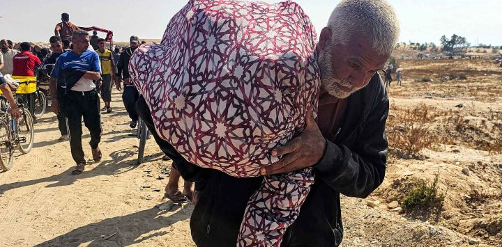 Un anciano camina con un saco al hombro mientras palestinos desplazados reciben ayuda humanitaria. Foto de AFP