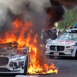 Manifestantes destrozan el parabrisas de un vehículo junto a un vehículo Waymo en llamas mientras se enfrentan con las fuerzas del orden en las calles que rodean el edificio federal durante una protesta tras los operativos federales de inmigración en Los Ángeles, California. | Foto:RINGO CHIU / AFP