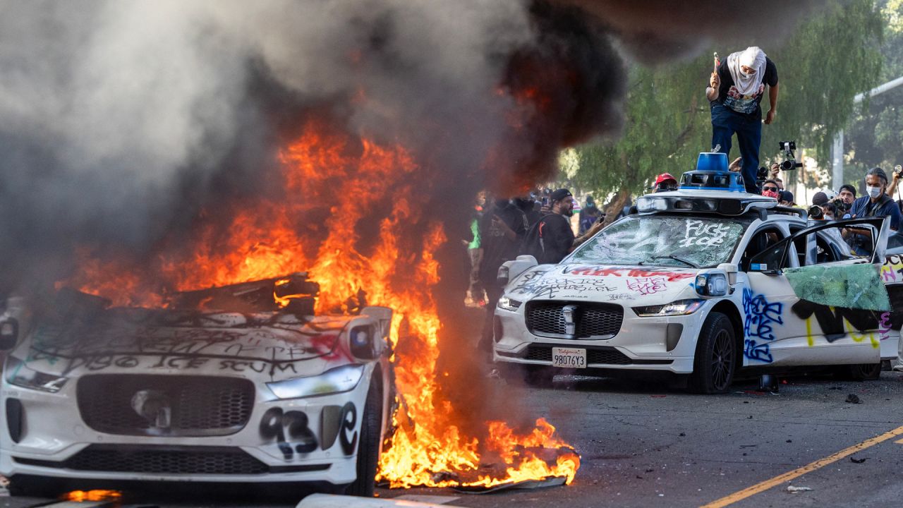 Manifestantes destrozan el parabrisas de un vehículo junto a un vehículo Waymo en llamas mientras se enfrentan con las fuerzas del orden en las calles que rodean el edificio federal durante una protesta tras los operativos federales de inmigración en Los Ángeles, California. | Foto:RINGO CHIU / AFP